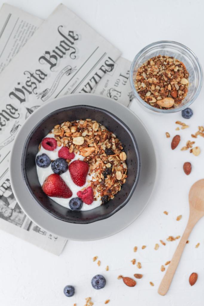 Bowl of granola, berries, and yoghurt, on top of a news paper and beside a smaller bowl of granola. 